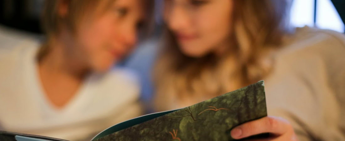 Imagem de uma menina acompanhada por uma mulher em frente a um livro de histórias, mostrando a importância dos hábitos e como uma rotina de estudo de inglês para crianças pode ajudar.