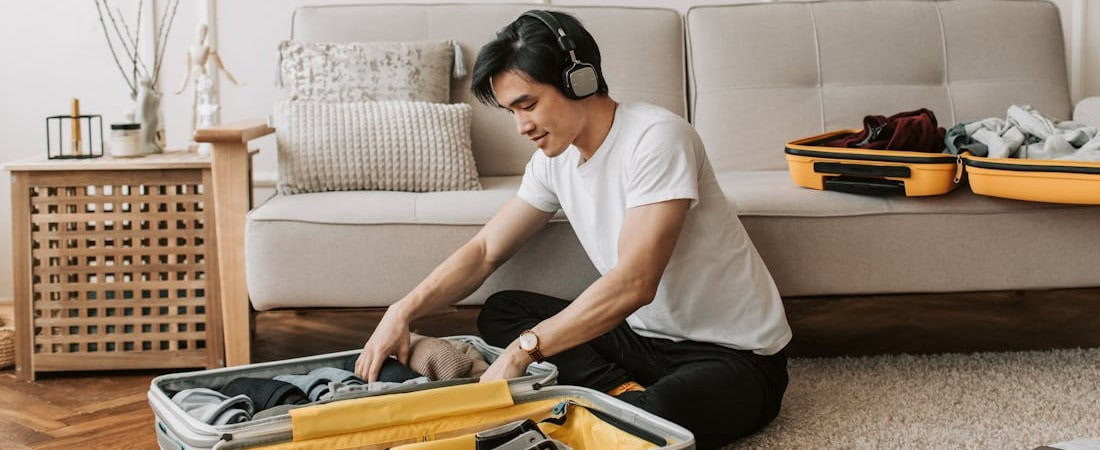 Imagem de um jovem asiático de camiseta branca e fones de ouvido pretos, sentado no chão da sala de estar e focado em fazer as malas, enquanto se prepara para estudar com o curso de Inglês Online para Viagens, cujo conteúdo está visível na tela de um notebook aberto dentro da mala.