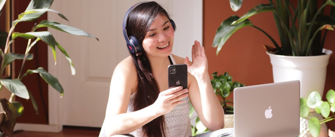 Imagem de uma mulher jovem e sonriente com fones de ouvido azuis, sentada no chão cercada por plantas enquanto acena com a mão diante de seu smartphone e notebook, refletindo como criar um ambiente de estudo divertido inglês em casa.