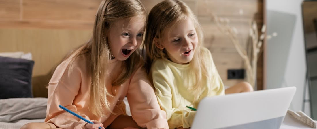 Imagem de duas meninas loiras com expressões de surpresa e alegria, sentadas em uma cama com lápis na mão enquanto olham fixamente para a tela de um notebook, mostrando como pode ser empolgante aprender inglês online divertido.