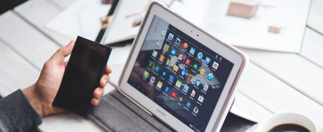 Imagem de uma mão segurando um smartphone ao lado de um tablet ligado com vários aplicativos na tela sobre uma mesa branca, ilustrando os diferentes recursos para aprender inglês rápido online.