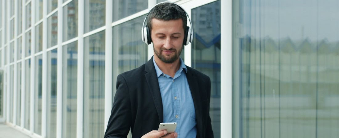 Imagem de um homem de terno preto e camisa azul caminhando em frente a um edifício espelhado, enquanto usa fones de ouvido e olha para o smartphone, sugerindo a praticidade de acompanhar aulas de inglês online eficazes em qualquer lugar.