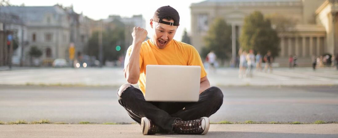 Imagem de um jovem de camiseta amarela e boné virado para trás, sentado de pernas cruzadas no chão ao ar livre e celebrando com entusiasmo diante de seu notebook, mostrando o sucesso obtido ao combinar videoaulas, jogos e exercícios de inglês.