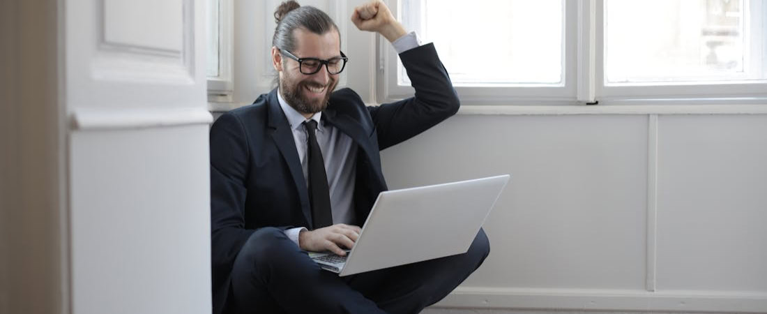 Imagem de um homem sentado em frente a uma parede com um laptop no colo, comemorando, referindo-se ao potencial deste blog quando os erros comuns ao aprender inglês online