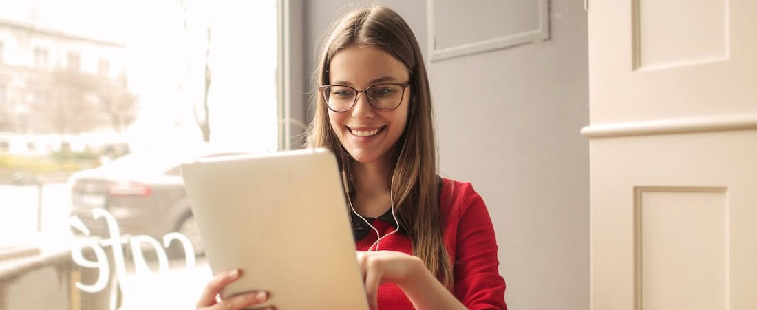 Imagem de uma jovem sorridente com óculos e blusa vermelha, sentada perto da janela de um café enquanto usa fones de ouvido e mexe em seu tablet, demonstrando tranquilidade ao escolher plataforma para aprender inglês.