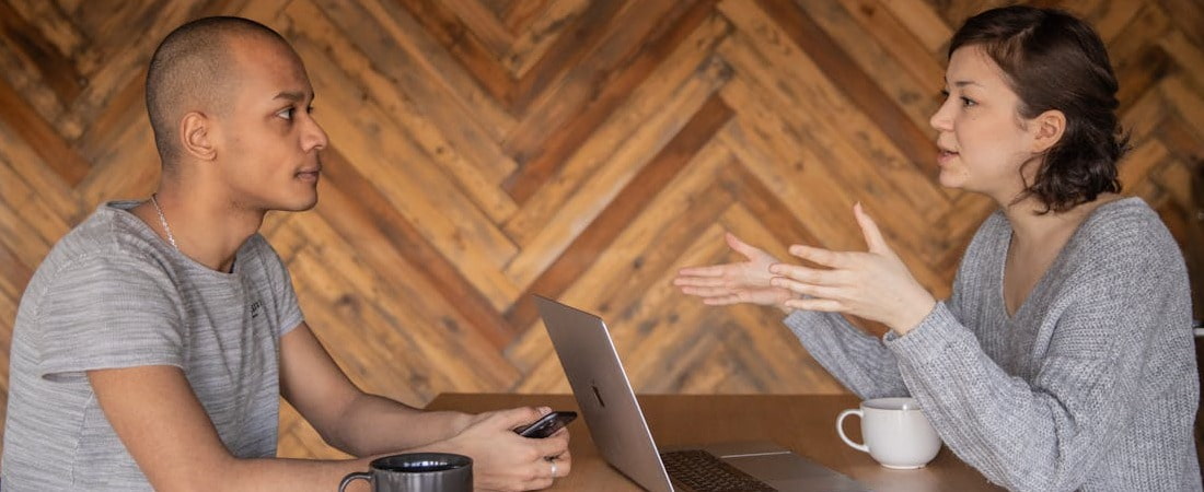 Imagem de um jovem e uma mulher sentados frente a frente em uma mesa de madeira com um notebook, engajados em uma conversa profissional onde ela gesticula ativamente, ilustrando a importância de dominar o inglês online carreira profissional.