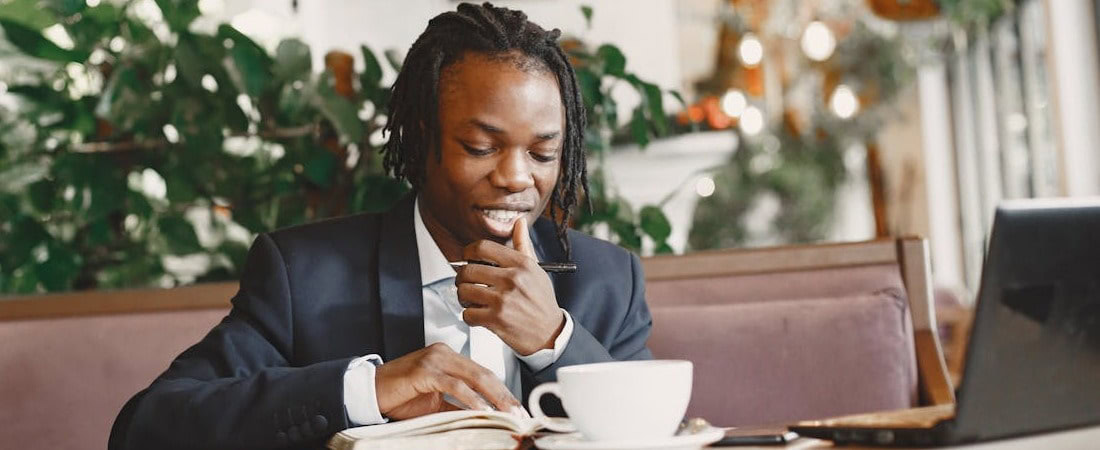 Imagem de um jovem profissional com dreadlocks usando terno, sentado em um café aconchegante enquanto analisa suas anotações com um sorriso leve, uma xícara de café e um notebook na mesa, mostrando foco para manter motivação aprender inglês online.