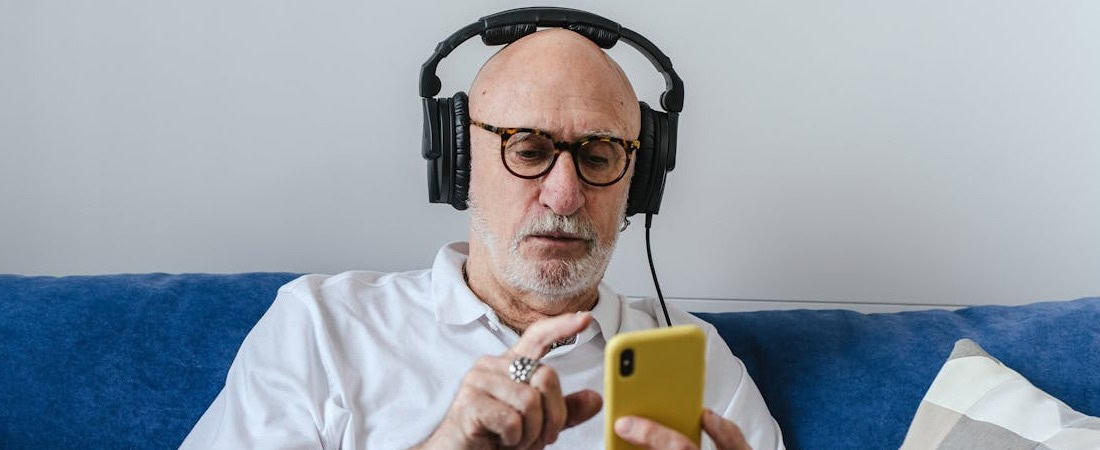 Imagem de um senhor idoso com óculos e fones de ouvido grandes, sentado em um sofá azul enquanto interage concentrado com o seu smartphone, aproveitando a tecnologia para melhorar listening inglês online.