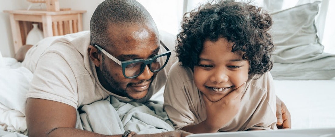Imagem de um homem com óculos e um menino sorridente com cabelo cacheado, ambos deitados em uma cama enquanto olham juntos para a tela de um dispositivo eletrônico, destacando a importância de motivar crianças aulas de inglês online.