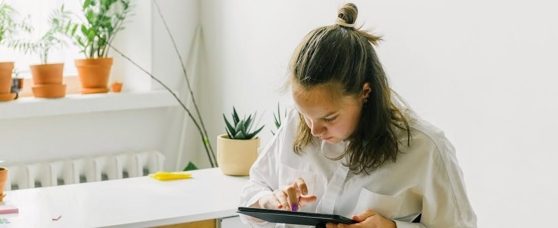 Imagem de uma jovem focada, sentada em uma mesa bem iluminada por luz natural e decorada com várias plantas, utilizando um tablet para praticar inglês online sem tempo, demostrando a flexibilidade de horários.