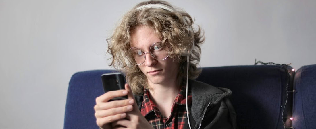 Imagem de um jovem de cabelos ondulados e óculos, vestindo uma camisa xadrez e fones de ouvido brancos, concentrado olhando para a tela de seu smartphone enquanto está sentado em um sofá azul, mostrando a praticidade de acessar recursos interativos inglês online.