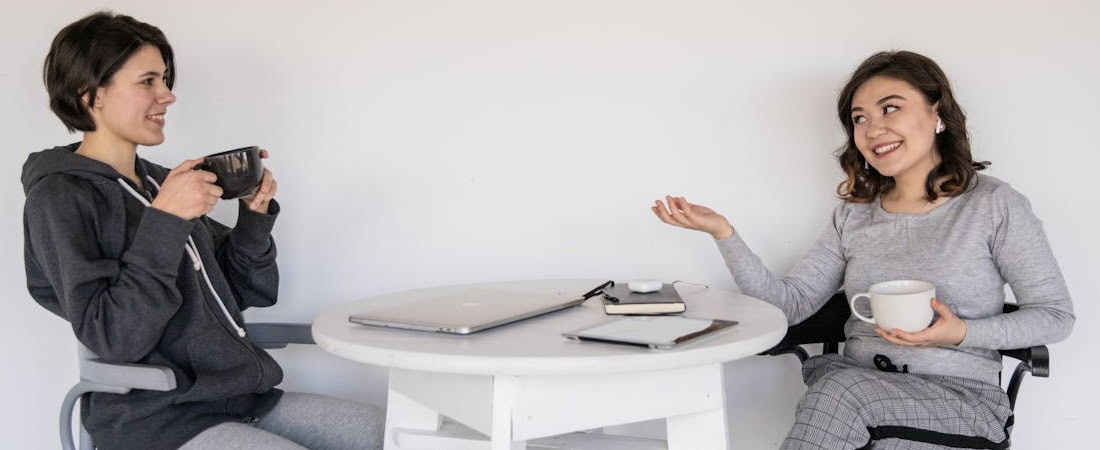 Imagem de duas amigas sorridentes sentadas ao redor de uma mesa redonda branca com canecas nas mãos, conversando de forma descontraída enquanto compartilham um espaço de estudos com notebook, excelente representação para quem deseja vencer timidez ao falar inglês.